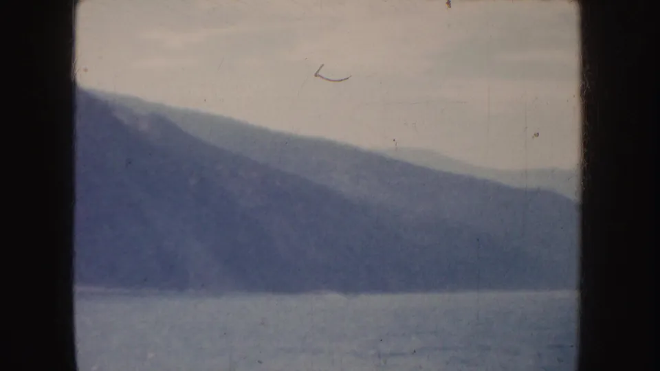 1952 California 8mm frame of a sloping mountain ridge over calm water, with muted color, dust and dark frame borders.