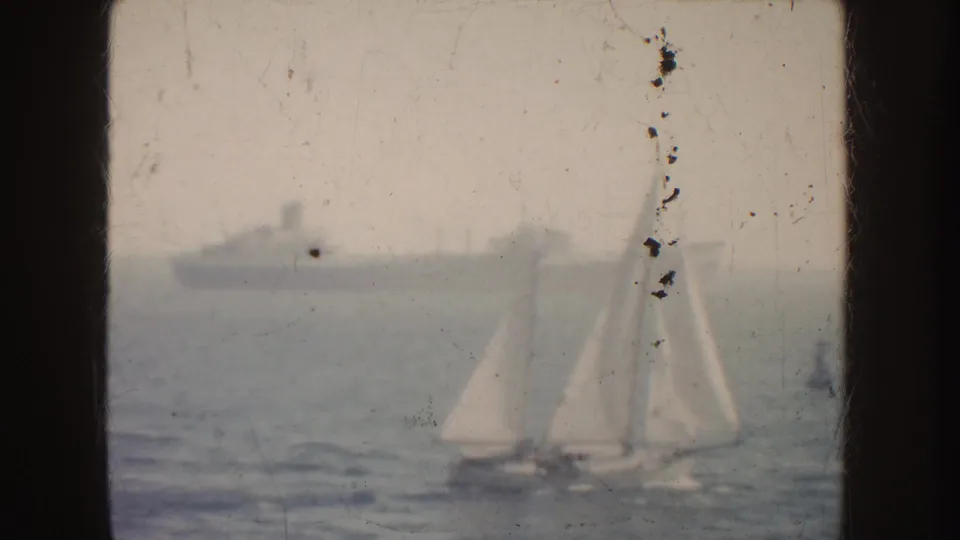 1952 California coastal home-movie frame with a white sailboat crossing hazy water and a large ship in the background.