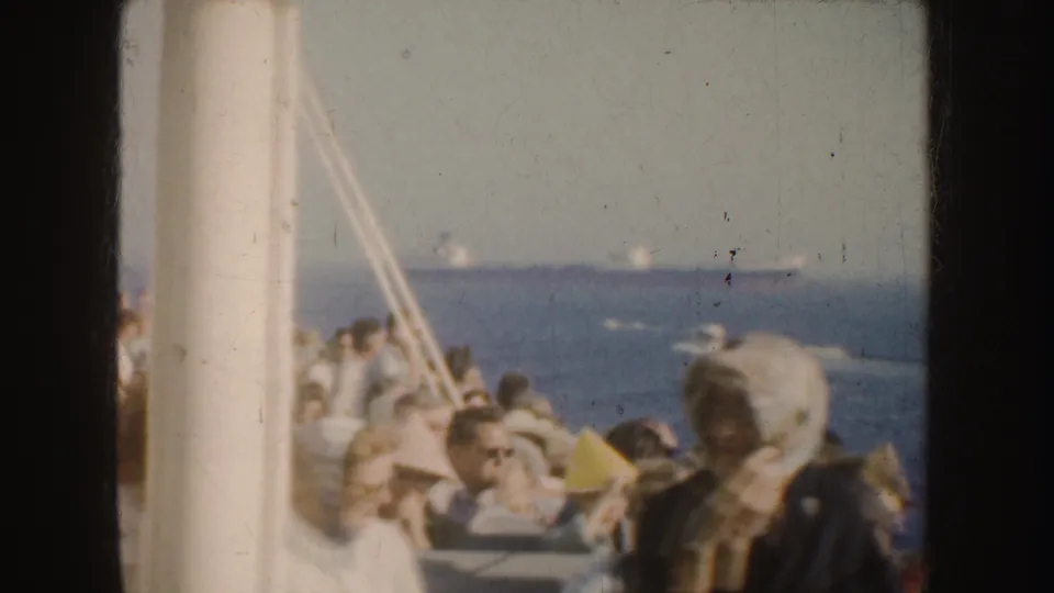 1952 California 8mm frame from a vessel deck, showing seated passengers with open water and distant ships in the background.