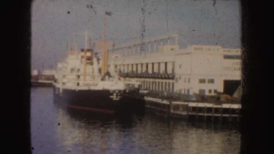 Soft 8mm view of a black-and-white ship moored beside a waterfront building and pier in California.