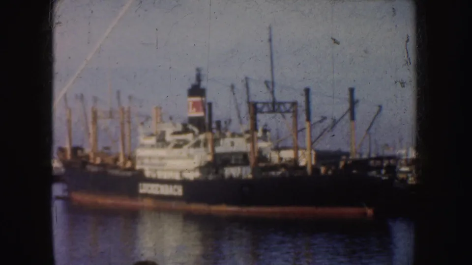 A dark-hulled ship with a funnel is moored near pilings and industrial dock structures in a 1952 California harbor scene.