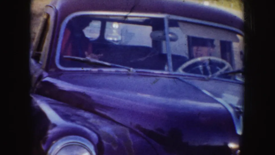 A mid-century car viewed from the front, with a person seated inside and rural ground behind it.