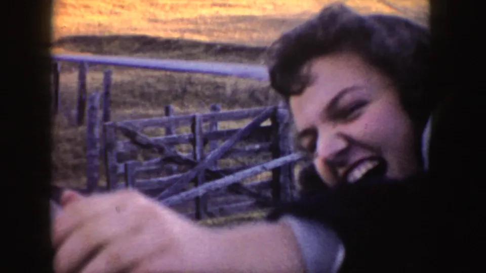 1955 Idaho home-movie frame of a smiling woman leaning from a car window toward a donkey beside rural fencing.
