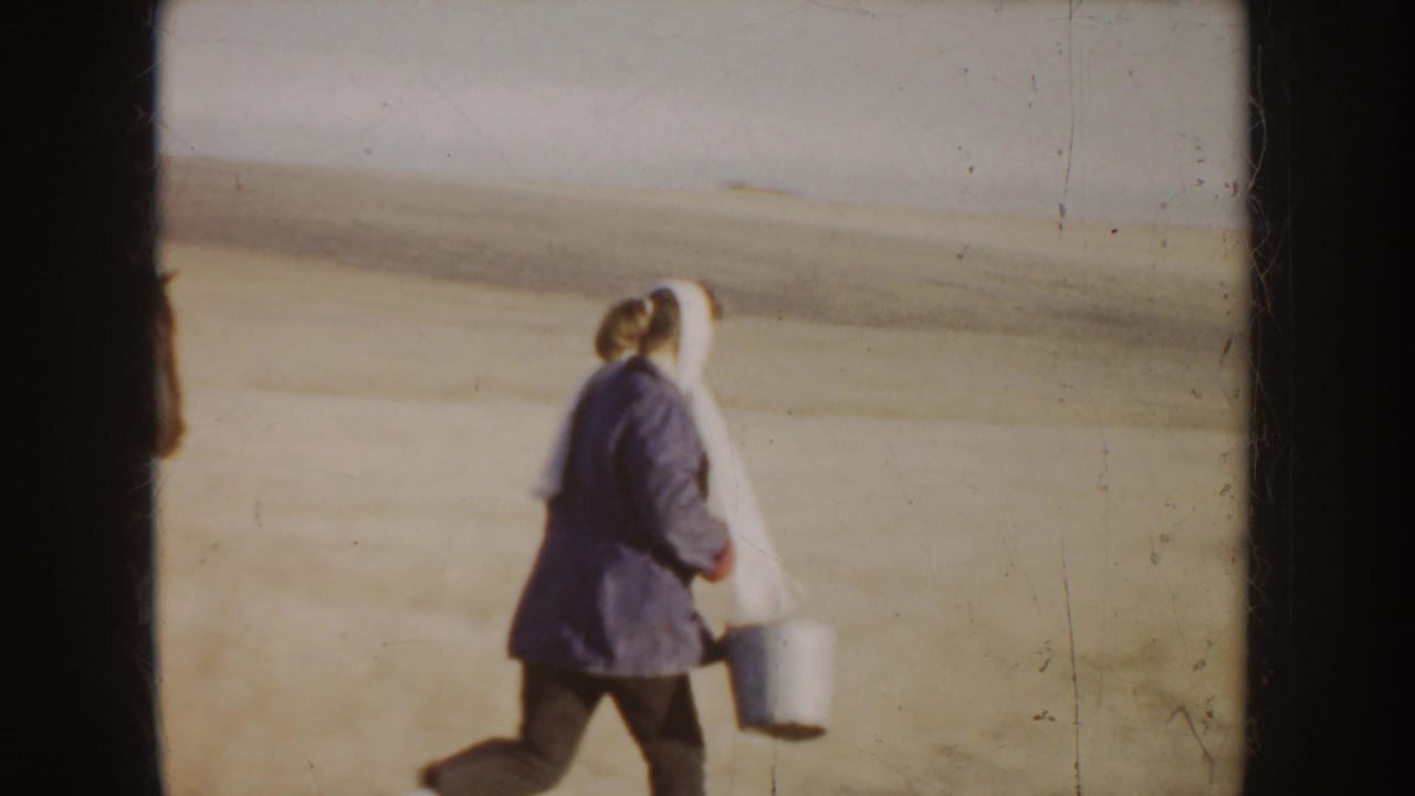 Woman in a coat and headscarf walks across a dry field carrying a metal bucket.
