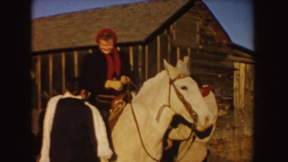 A person wearing red sits on a light horse near a rustic wooden barn in Idaho, with another figure partly visible in the foreground.