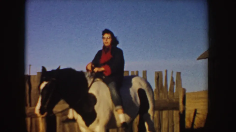 A rider in dark clothing and red scarf sits on a dark-and-white horse near a rough wooden fence under clear sky in Idaho.