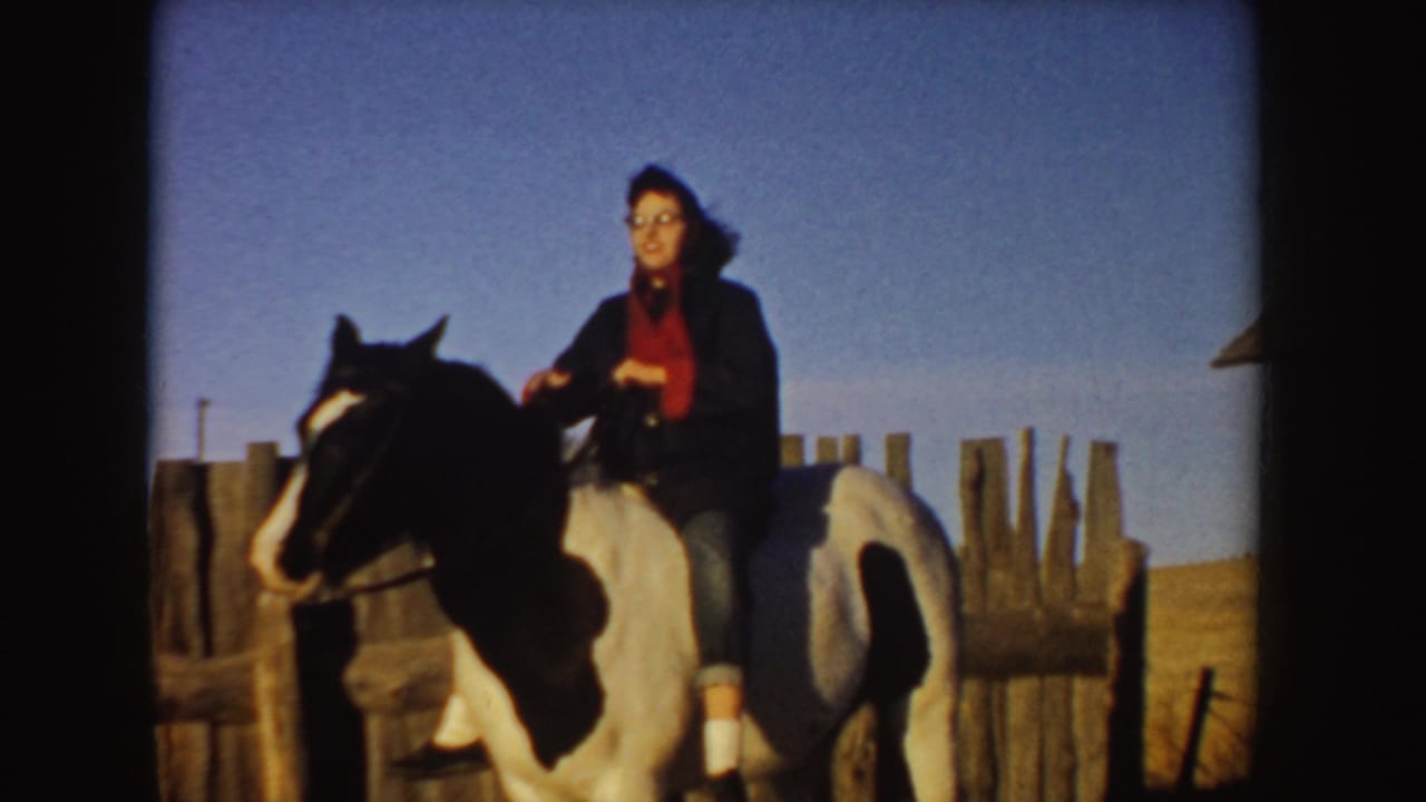 In supported 1955 Idaho footage, a woman rides a black-and-white horse near rough wooden fencing in open countryside.
