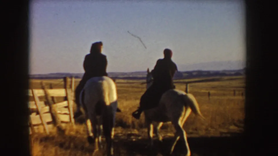 Two riders on light-colored horses head away from the camera through a fence opening in the Idaho prairie setting.