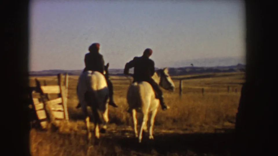 The riders continue away from the camera on light horses, framed by a gate and wide grassland in the supplied Idaho clip.