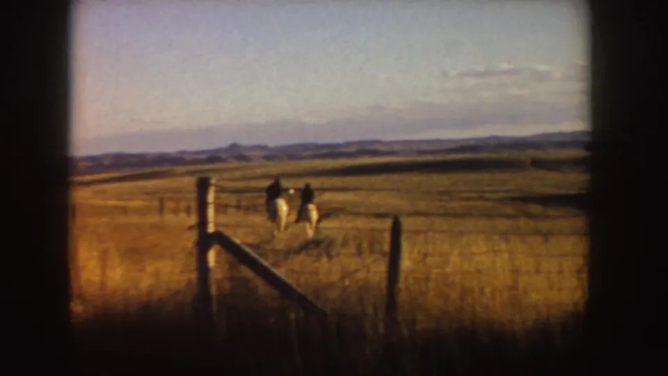 Two small horseback figures ride across a broad Idaho field, with fence posts, dry grass, and low hills in the distance.