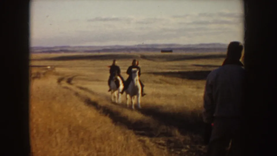 Two men ride white horses in an Idaho meadow as a third man stands near the camera at the side of the frame.
