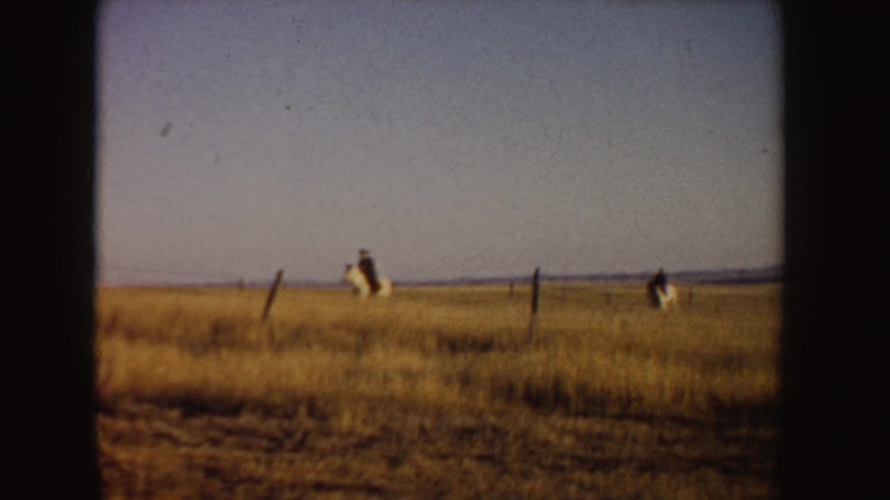 Wide 1955 Idaho home-movie frame showing distant horseback riders in golden prairie grass near simple fence posts.
