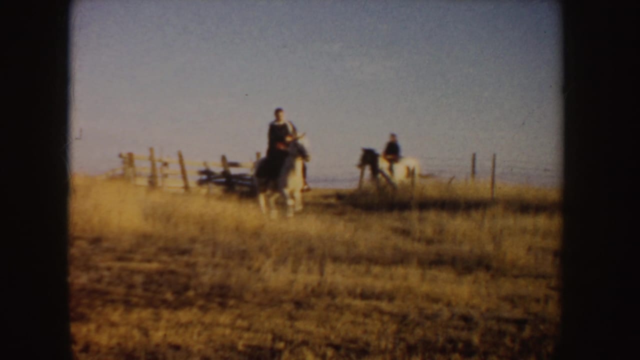 Two riders on horses near a wooden fence in open grassland.