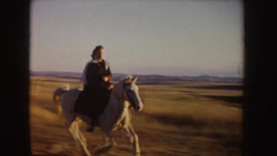 Closer 1955 Idaho home-movie view of a woman riding a light horse across golden prairie with low hills in the distance.
