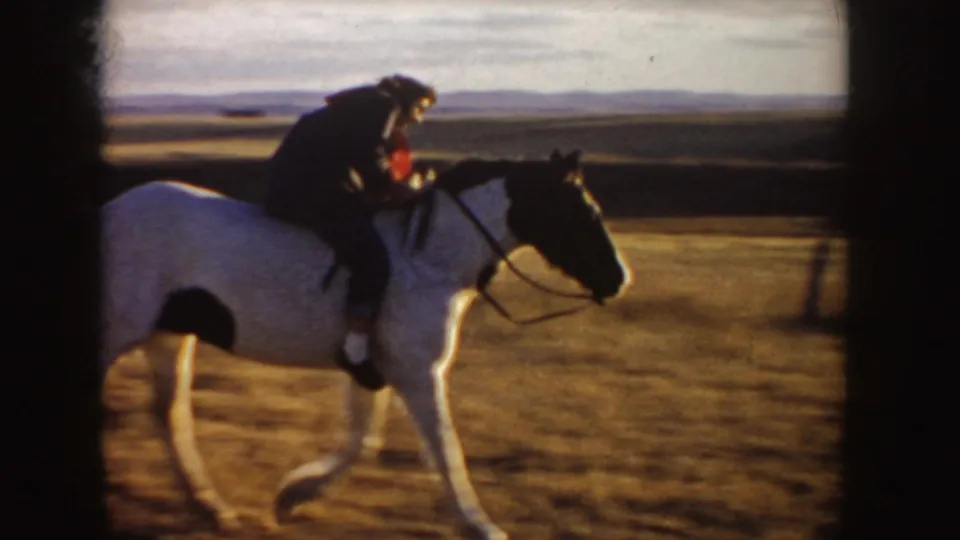 1955 Idaho home-movie frame of a rider leaning forward on a white horse against a broad plains horizon.