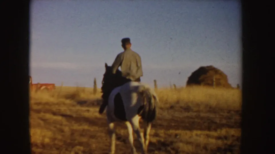 A man rides a white horse away from the camera across an Idaho field, with fence posts and a low hay-like mound in the distance.