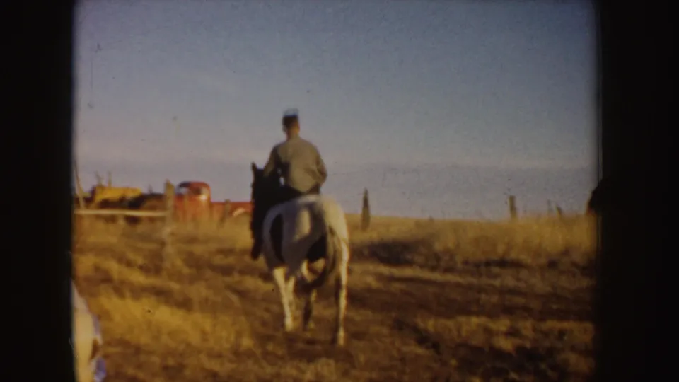 The rider continues away on the white horse through dry grass, with a red vehicle or equipment shape visible near the horizon.