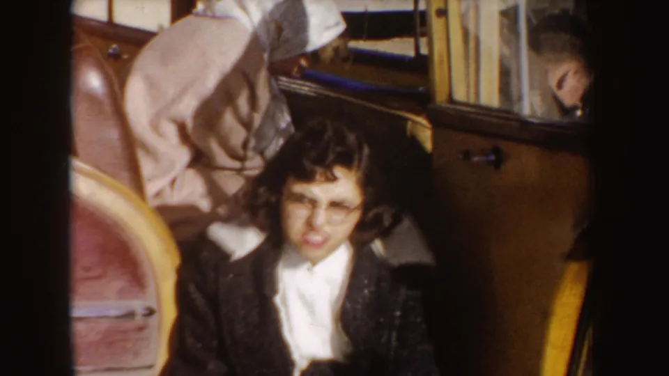 A woman sits inside a vehicle interior, surrounded by other passengers and luggage or upholstery in 1950s home-movie footage.