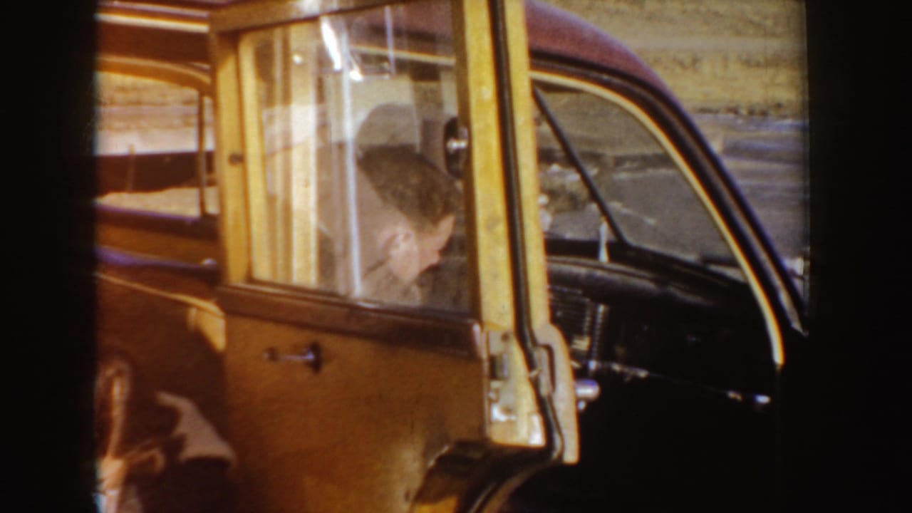 1955 Idaho home-movie frame with a man seated at the wheel of a classic car, seen through the side window and open doorway.