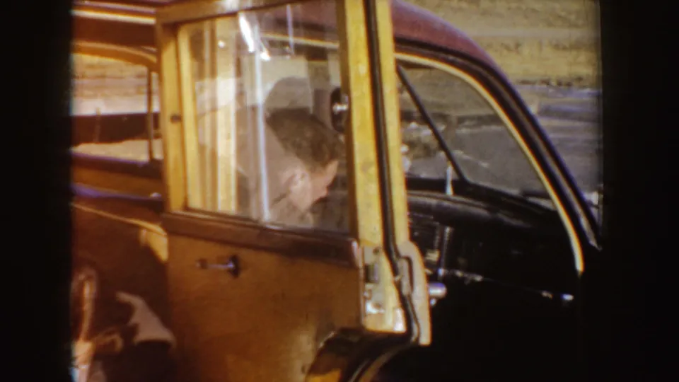 1955 Idaho home-movie frame with a man seated at the wheel of a classic car, seen through the side window and open doorway.