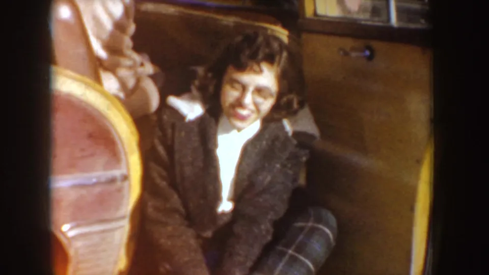 1955 Idaho 8mm frame showing a smiling woman with glasses, dark jacket, and plaid pants seated in a classic car interior.