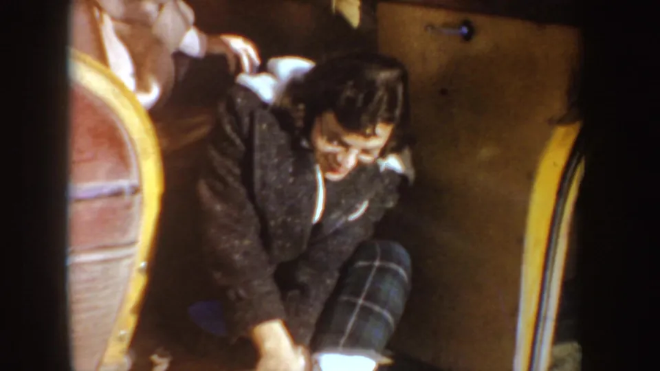 1955 Idaho home-movie frame of a woman in glasses and plaid pants adjusting her position in the back seat of a classic car.