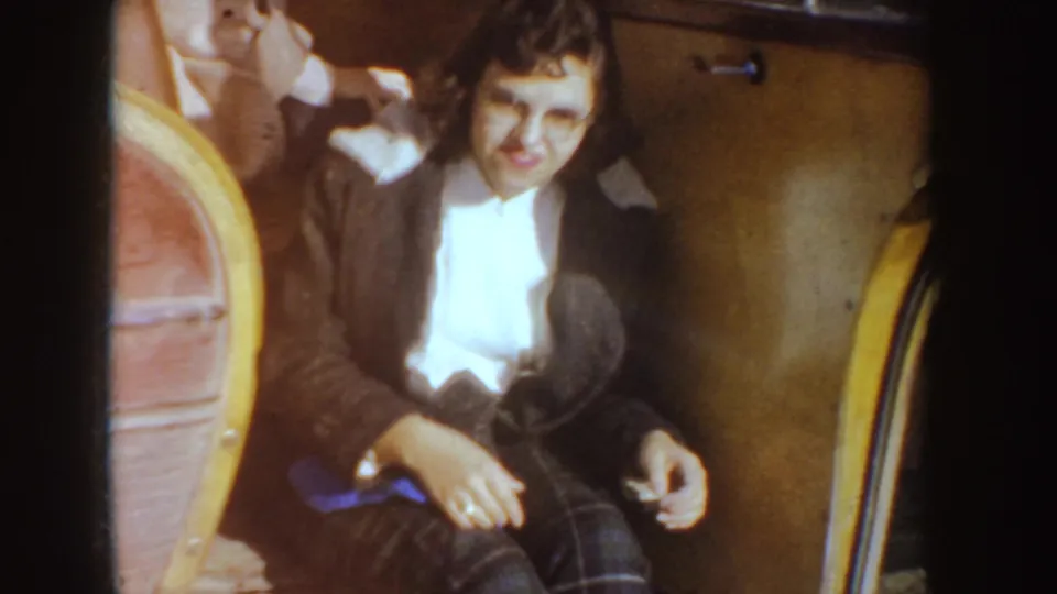 A smiling woman in a dark coat sits inside a vintage car, with the rounded seat and door panel visible in the warm 8mm frame.