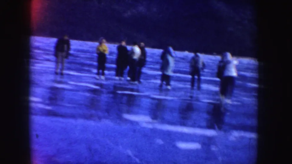 People in winter coats skate on a frozen Idaho lake, while a blurred skater passes in the foreground against the dark shoreline.
