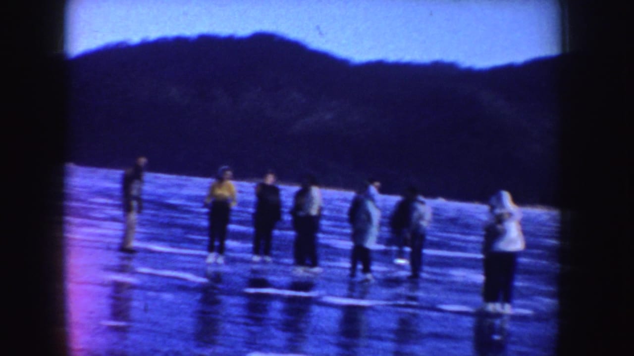 Several people stand and skate on a frozen lake with dark hills behind them.