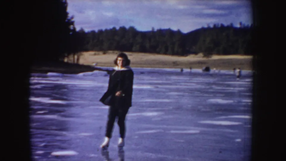 1955 Idaho 8mm still of a woman in a dark winter coat skating on a frozen lake before a wooded shoreline.