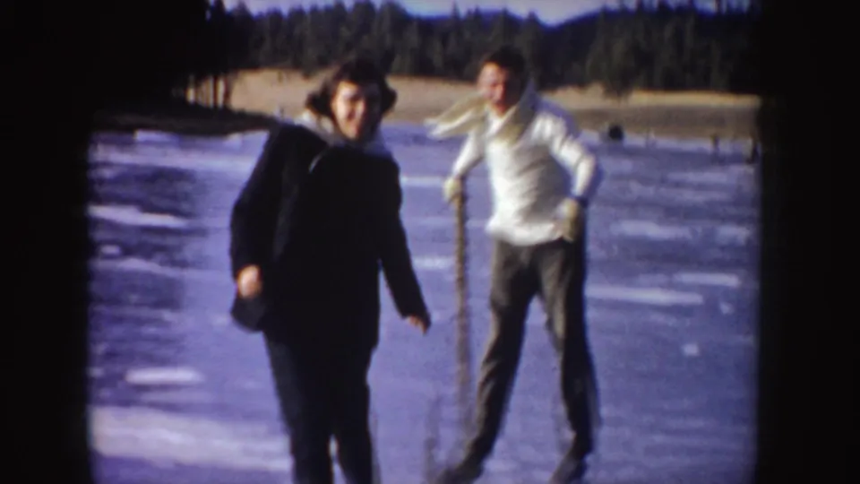 1955 Idaho home-movie frame showing a woman and a man skating toward the camera on a frozen lake, with trees along the shore.