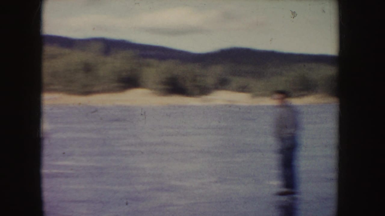 In 1955 Idaho home-movie footage, a blurred skater moves across a frozen pond or lake with trees and low hills in the background.