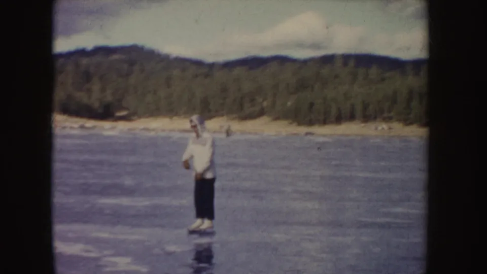 A person in a white jacket and dark pants pauses on skates on a frozen pond or lake in metadata-supported 1955 Idaho.