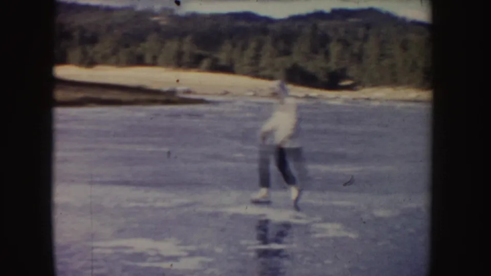 A light-jacketed skater glides across reflective ice in a 1955 Idaho home-movie frame with forest and hills beyond.