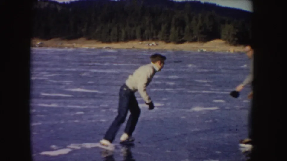 A skater in a white jacket leans into a turn on frozen water, with another skater partly visible at frame edge in 1955 Idaho footage.