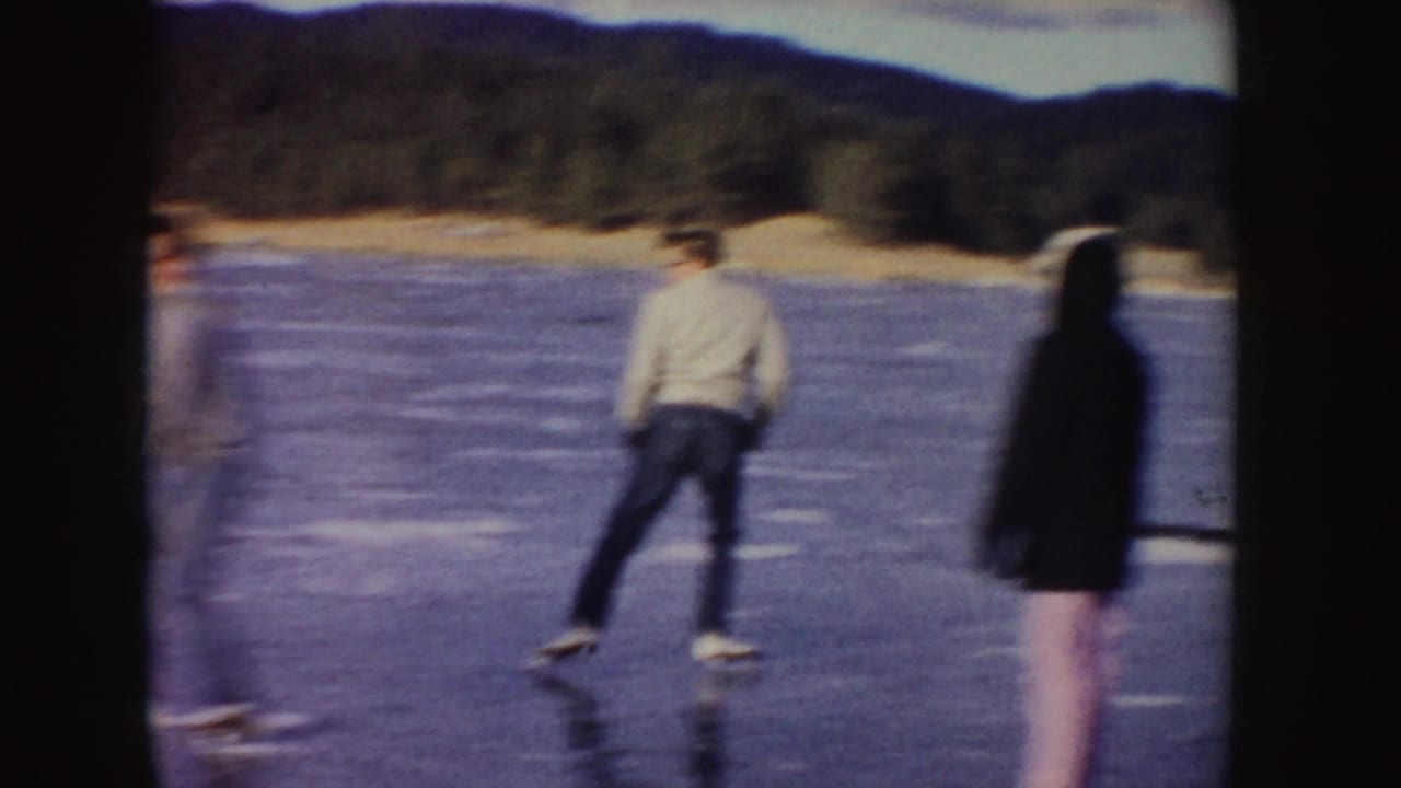 Two people skating on a frozen lake with forested hills in the background.