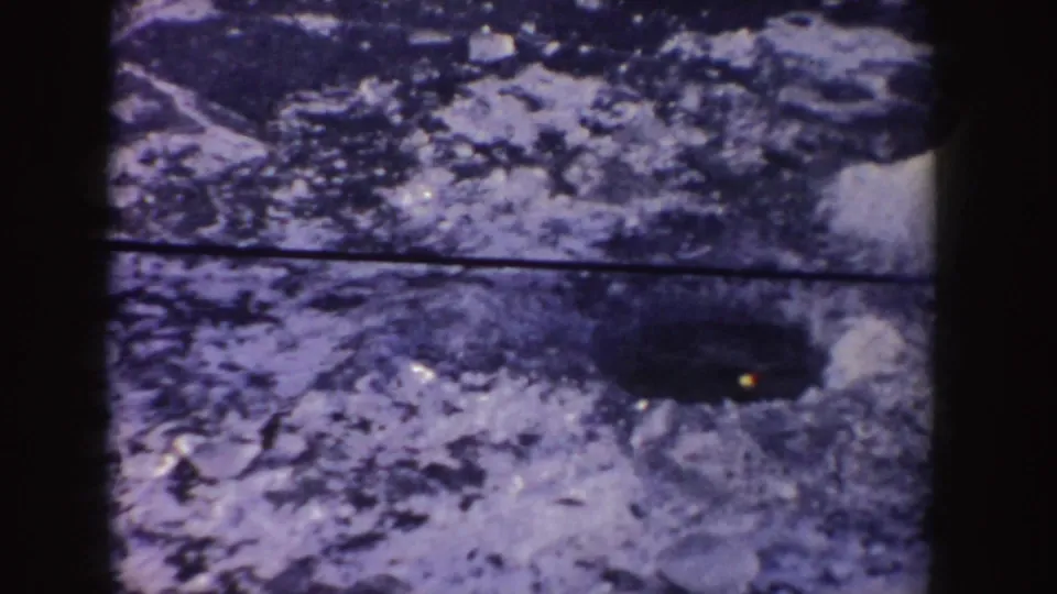 1955 Idaho home-movie frame showing a dark hole in snow or ice with a small yellow light near its edge.