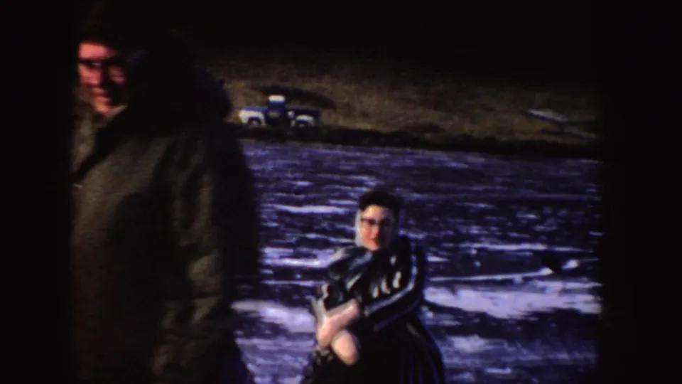 1955 Idaho 8mm frame showing two visible skaters on a frozen lake, with a parked vehicle and shoreline in the background.