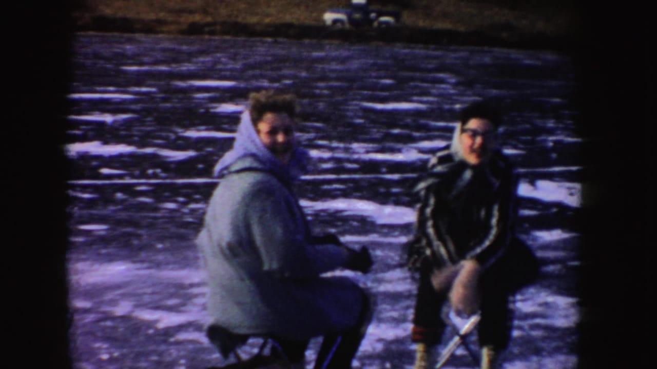 Two people crouch or sit on the frozen lake while looking toward the camera.