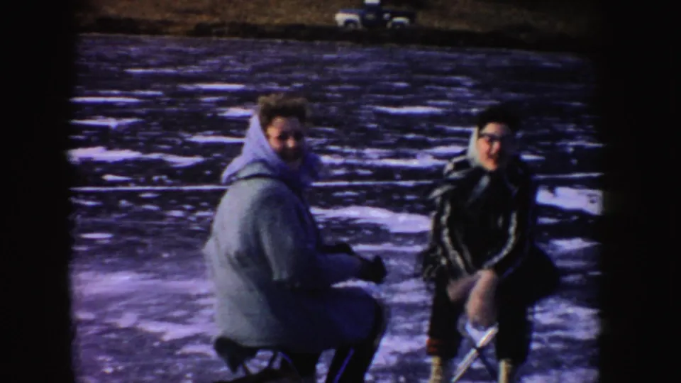 1955 Idaho 8mm frame with two skaters crouched on the ice, dressed in winter coats against the frozen lake surface.