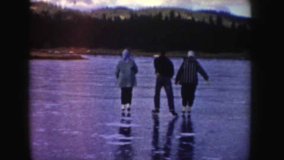 1955 Idaho 8mm frame showing three skaters from behind moving across a frozen lake with wooded hills beyond.