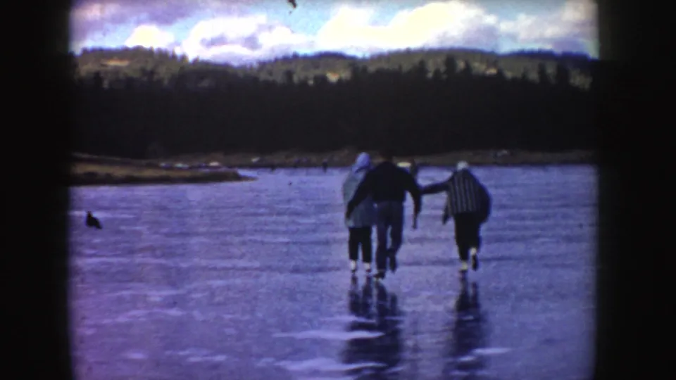1955 Idaho 8mm frame of three skaters receding across a frozen lake, backed by shoreline, trees, and winter sky.