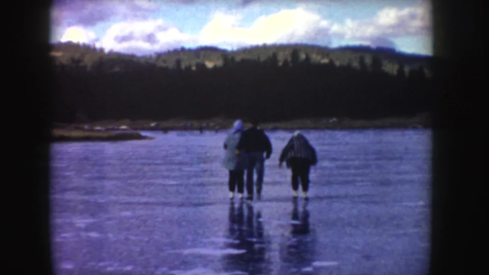 1955 Idaho home-movie frame showing four bundled people on a frozen pond beneath a cloudy sky and dark treeline.