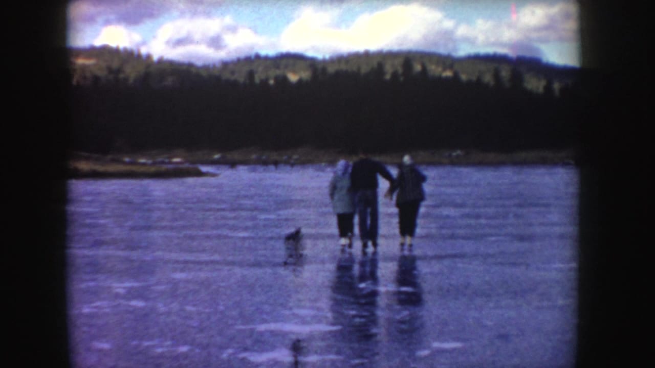 1955 Idaho frame with three bundled people on a frozen pond, a small dog-like figure nearby, and forested hills beyond.