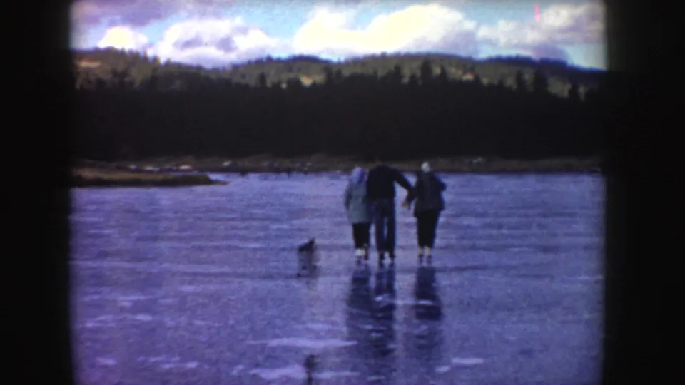 1955 Idaho frame with three bundled people on a frozen pond, a small dog-like figure nearby, and forested hills beyond.