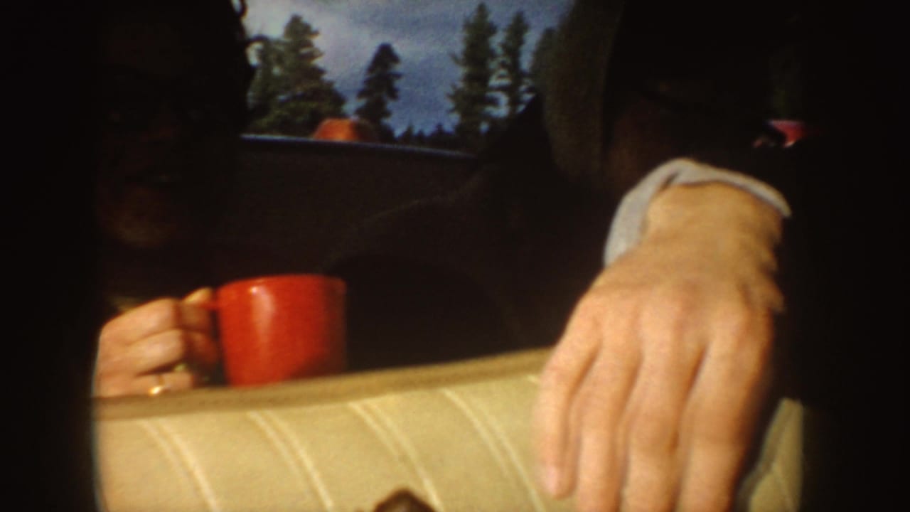 1955 Idaho home-movie frame with a passenger holding a red coffee mug inside a car, with trees visible through the window.