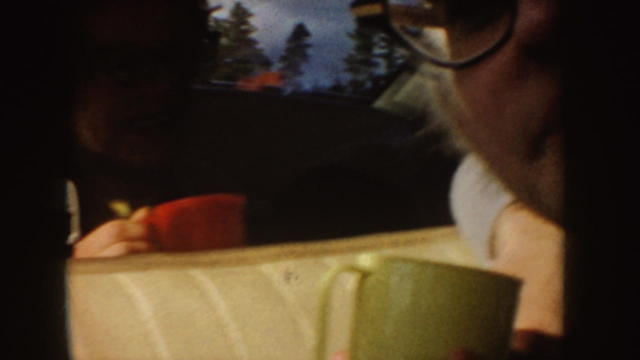 1955 Idaho home-movie frame showing passengers inside a car, including a person with glasses, with red and green mugs in the foreground.