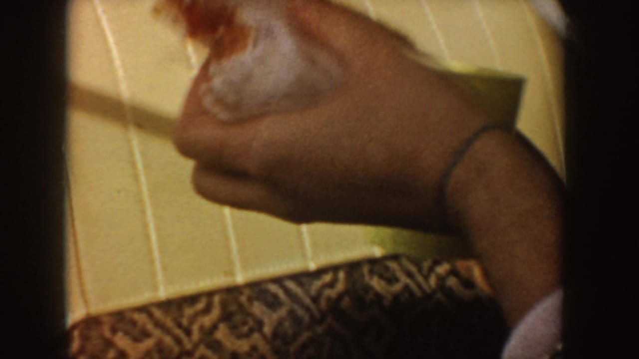 1955 Idaho home-movie close-up of a passenger hand holding a sandwich inside a car, with patterned clothing and cream upholstery visible.
