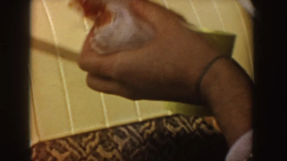 1955 Idaho home-movie close-up of a passenger hand holding a sandwich inside a car, with patterned clothing and cream upholstery visible.