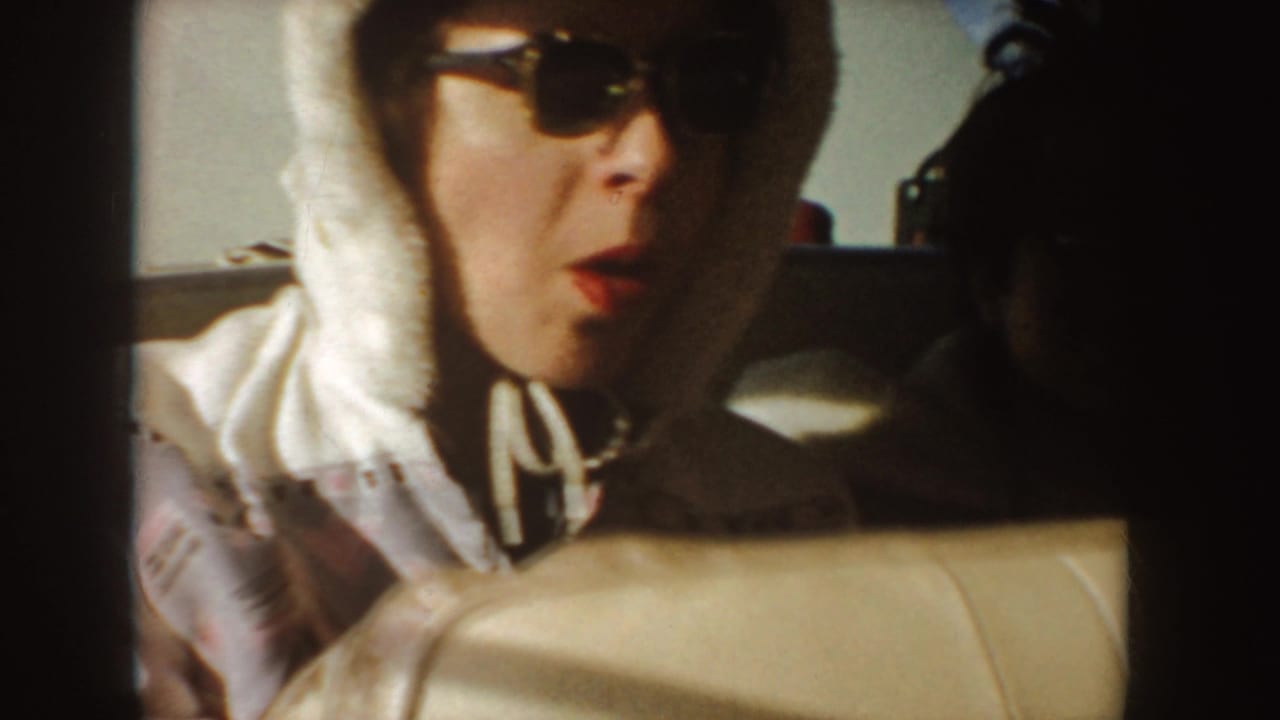 Woman in sunglasses and a white hooded jacket sits behind a car seat.